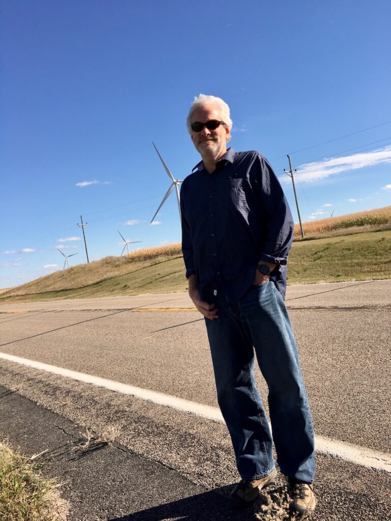 Writer, journalist, and podcaster Tom Schueneman stands by the side of a road in North Dakota with wind turbines in the background